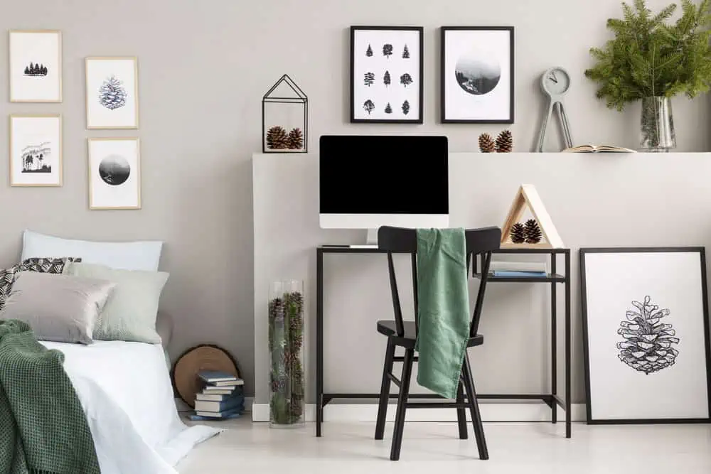 Bedroom with green cloth on black wooden chair at desk with computer, wooden triangle and cones, real photo with gallery of posters on empty grey wall